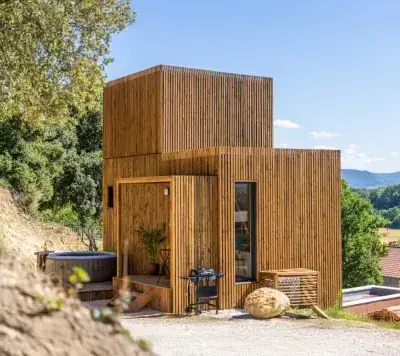 Studio de jardin Greenkub à bardage bois vertical, volume à étage et porte vitrée, posé sur un terrain en pente face à la campagne