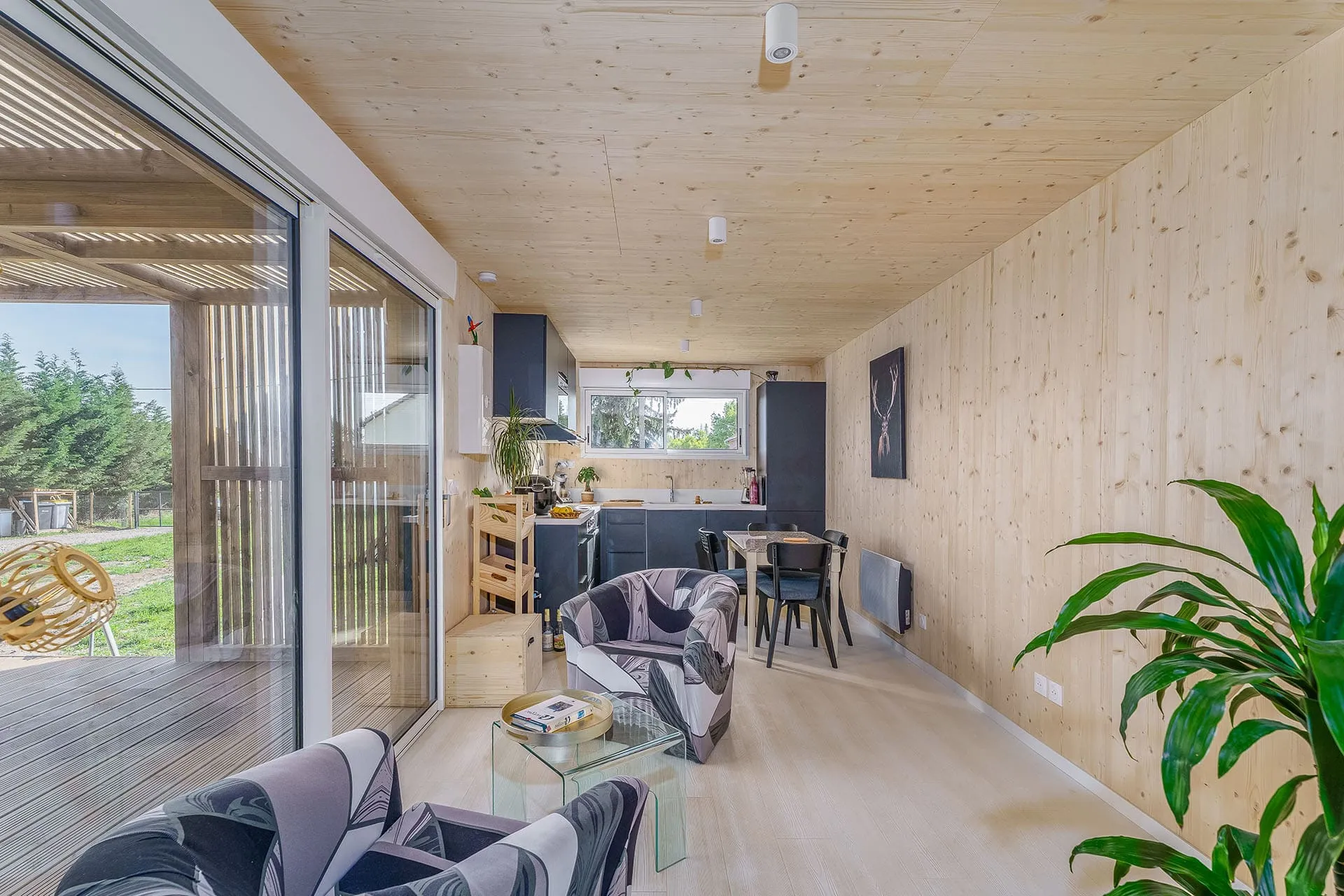 Intérieur salon-cuisine d’une maison bois Greenkub GK50 · murs et plafond en bois massif épicéa naturel, cuisine en L anthracite, baie vitrée coulissante ouvrant sur terrasse bois