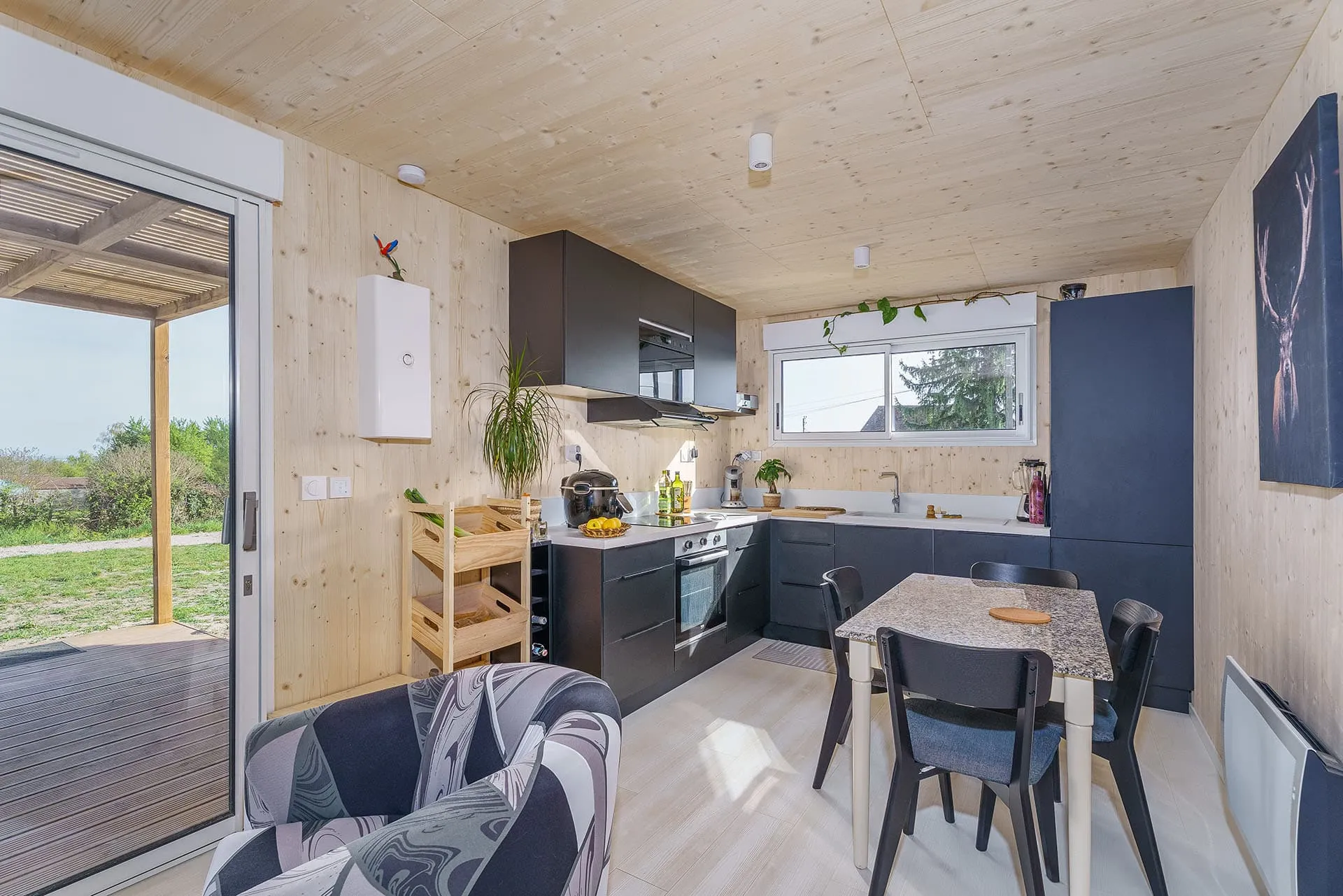Intérieur maison ossature bois Greenkub · séjour lumineux avec baie vitrée, cuisine équipée noire et murs bois naturel