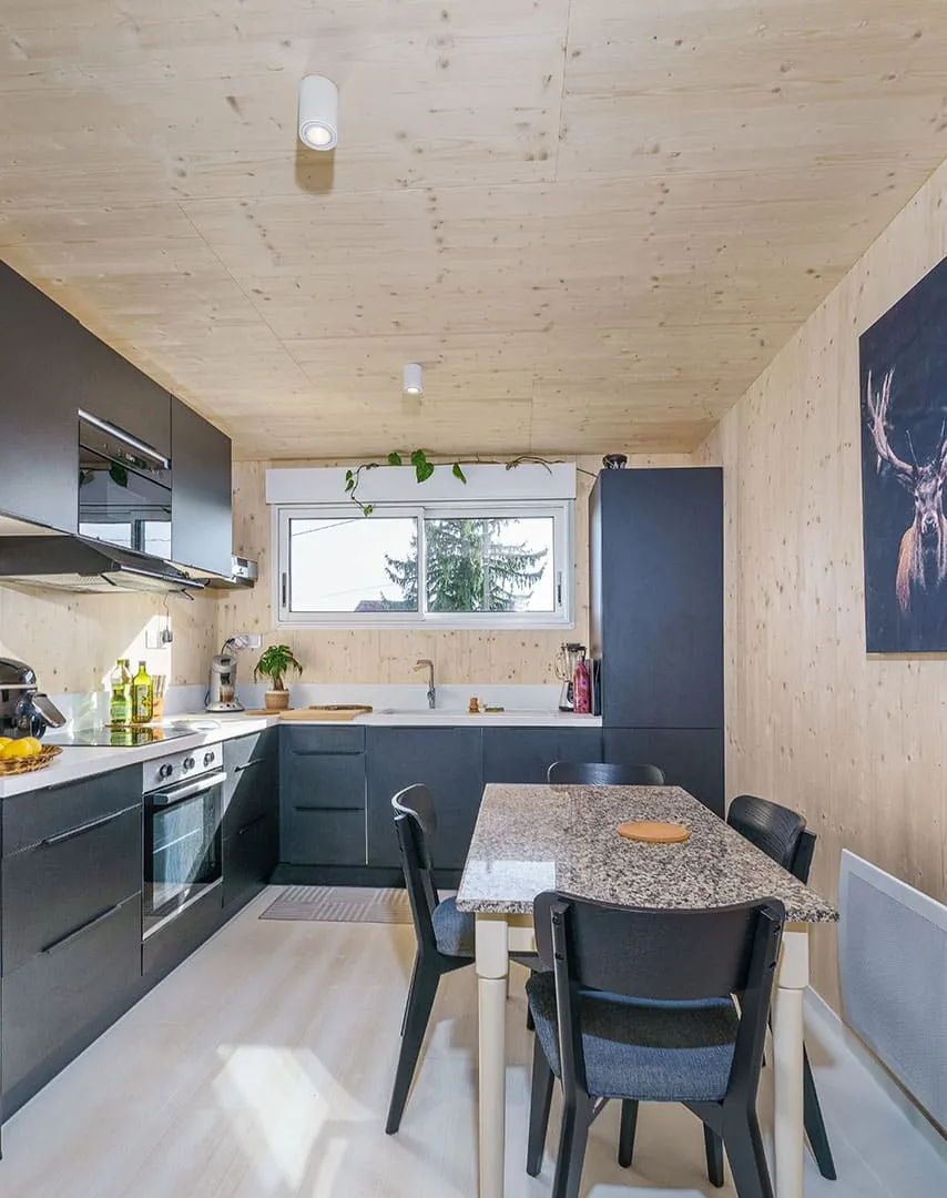 Cuisine équipée anthracite d’une maison bois Greenkub GK50 · murs bois massif épicéa, plan de travail blanc, table de repas avec plateau granit, spots plafond cylindriques blancs