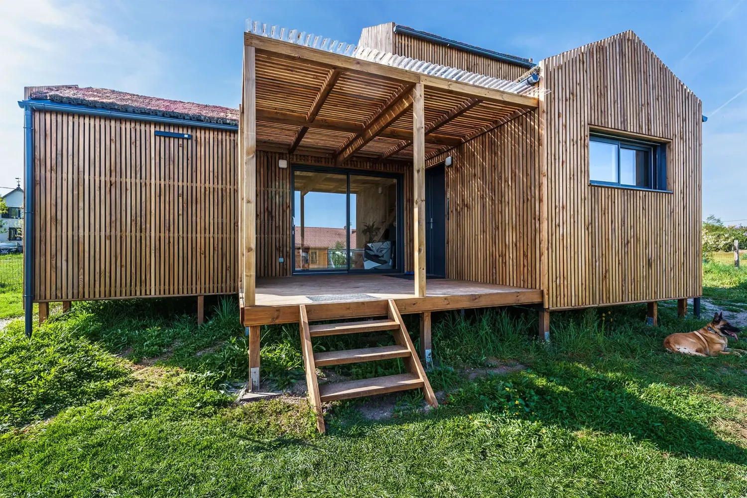 Façade principale d’une maison bois Greenkub GK50 · bardage bois vertical, toiture végétalisée multi-volumes, terrasse couverte avec pergola, structure sur plots métalliques