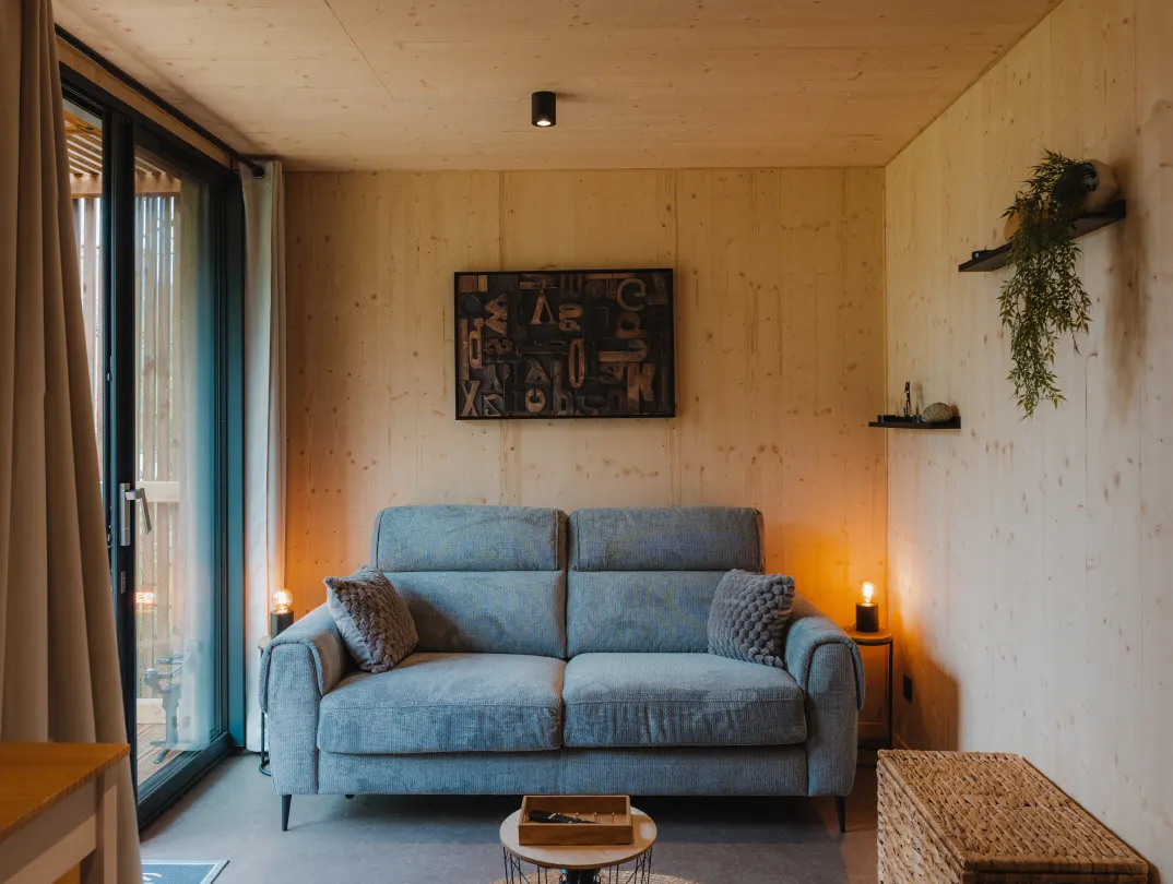 Intérieur d’un studio de jardin : coin séjour avec canapé, parement intérieur en bois massif épicéa et baie vitrée sur la terrasse